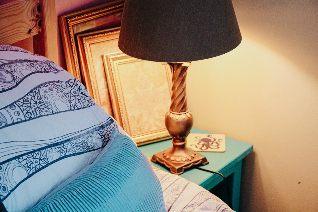 Edge of bed with blue cushion, next to blue bedside table with lamp and framed pictures in background.